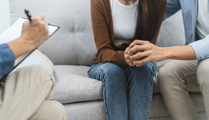 Una mujer sentada en un sofá, recibiendo apoyo emocional de un hombre mientras un terapeuta toma notas en una libreta. La escena muestra un ambiente de terapia y cuidado psicológico.