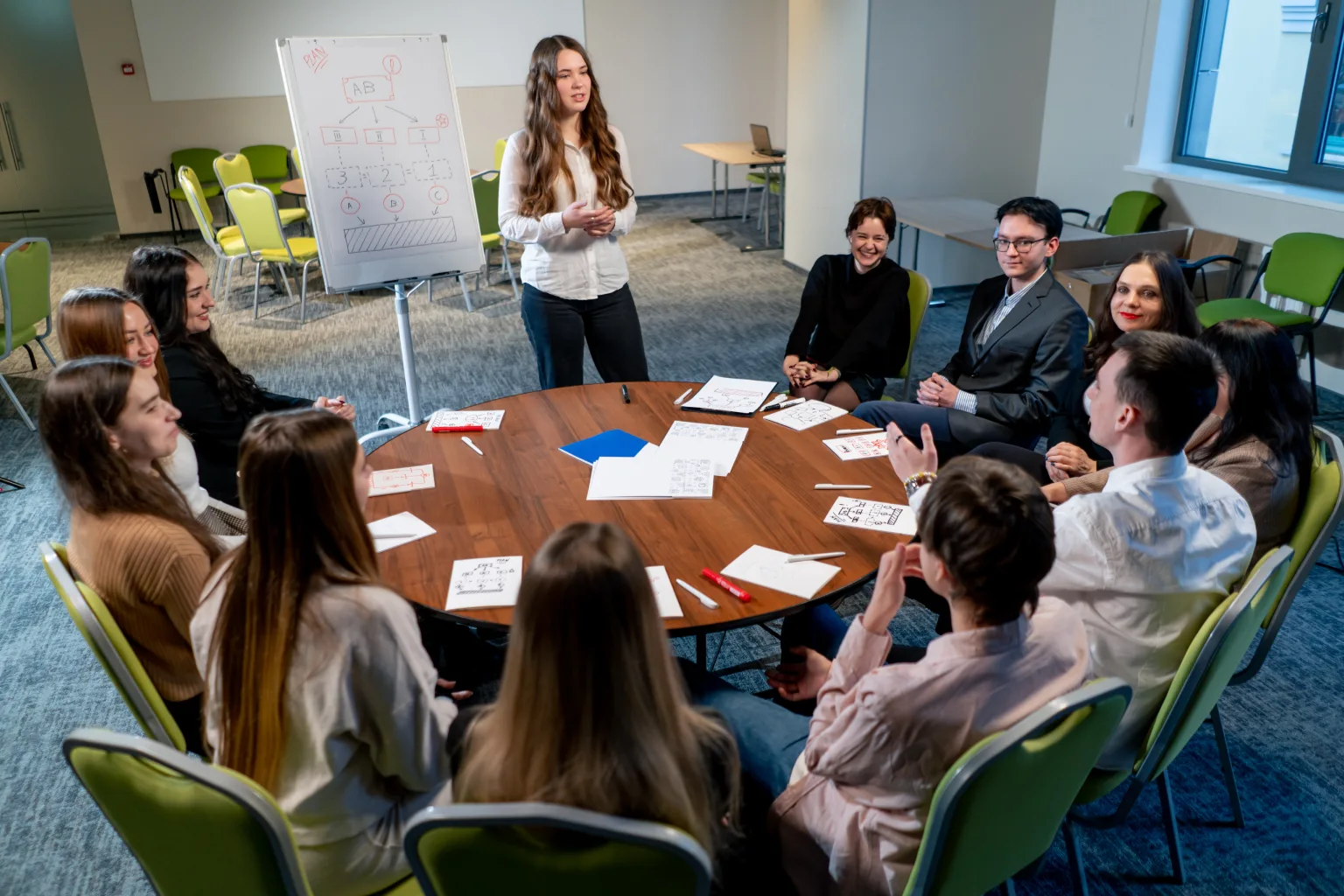 Reunión de equipo en una sala de conferencias, donde una mujer presenta ideas frente a un grupo diverso de personas sentadas alrededor de una mesa. Se observan papeles y materiales de trabajo, destacando un ambiente colaborativo y creativo.