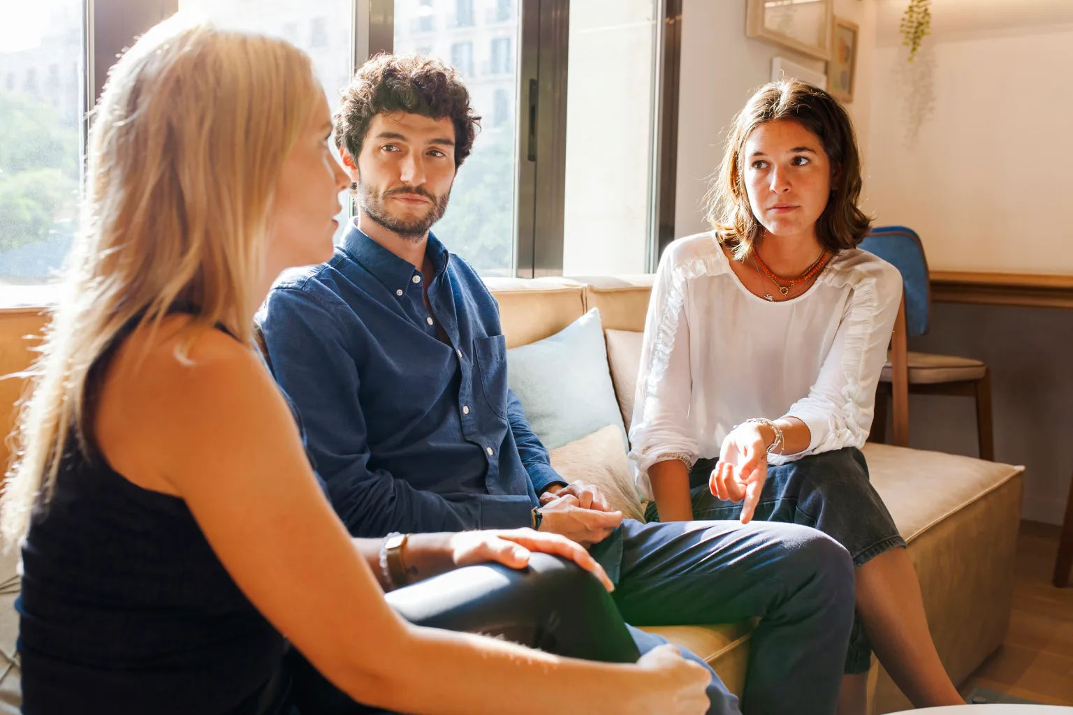 Tres personas conversando en un ambiente acogedor, con luz natural. Dos mujeres y un hombre sentados en un sofá, mostrando interés en la conversación. La mujer a la izquierda tiene el cabello largo y rubio, mientras que la mujer a la derecha tiene el cabello corto y oscuro. El hombre en el centro lleva una camisa azul y parece reflexionar sobre el tema discutido.