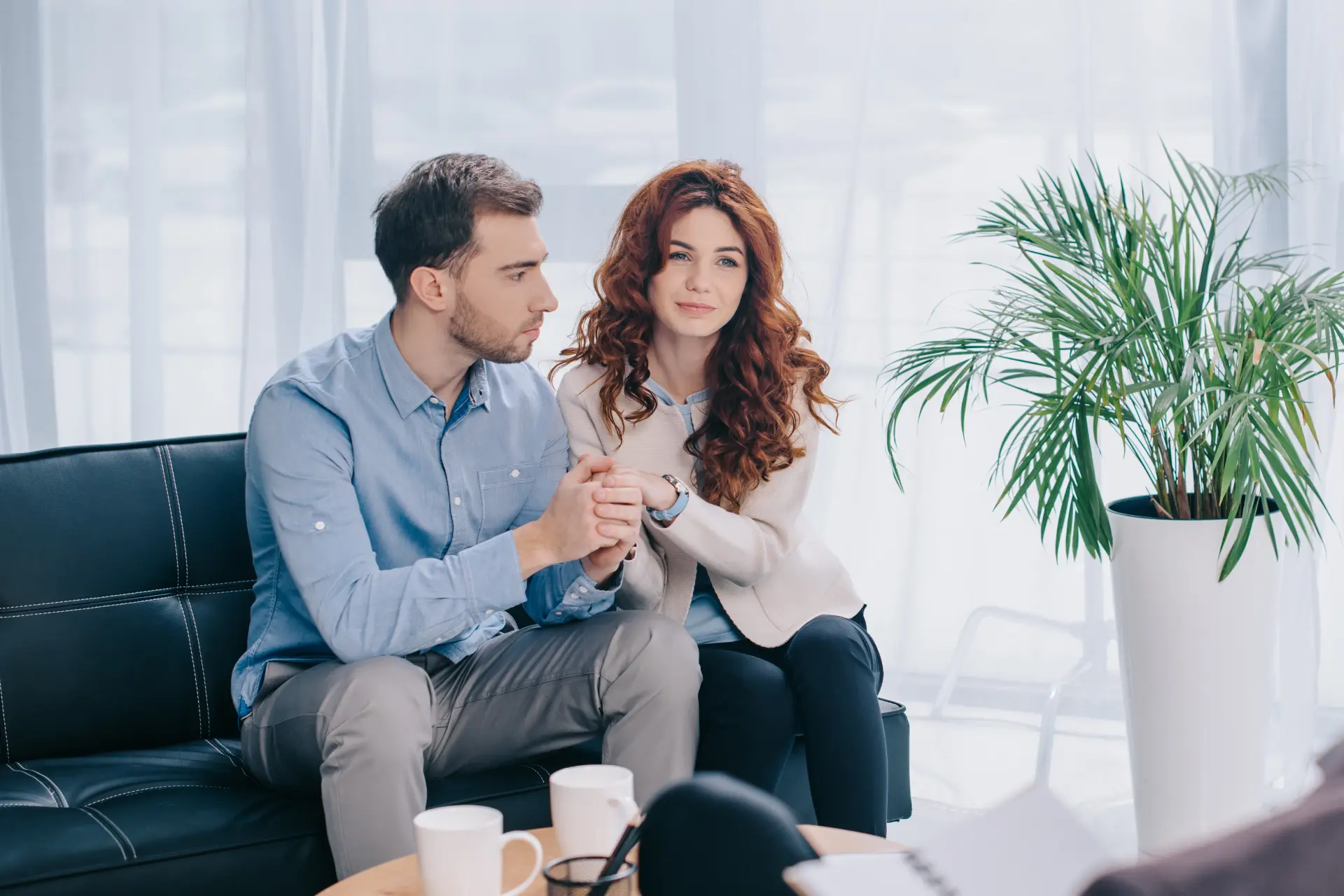 Pareja sentada en un sofá, mostrando apoyo emocional, mientras sostienen sus manos. En la mesa hay tazas de café y un cuaderno, sugiriendo una sesión de terapia o asesoramiento en un ambiente acogedor y luminoso.